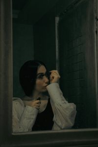 A woman is seen through a mirror, applying mascara in a dimly lit bathroom. She wears a loose white blouse over a black top, her expression calm and introspective. The muted lighting and slight grain give the image a moody, reflective atmosphere.